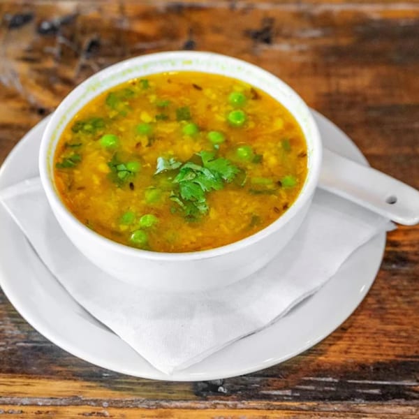 A white bowl of yellow Indian curry with green peas and cilantro, on a white plate with a spoon.