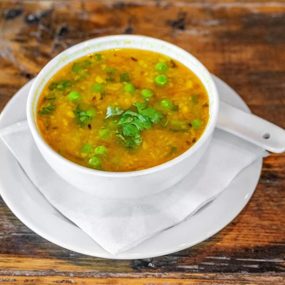 A white bowl of yellow Indian curry with green peas and cilantro, on a white plate with a spoon.