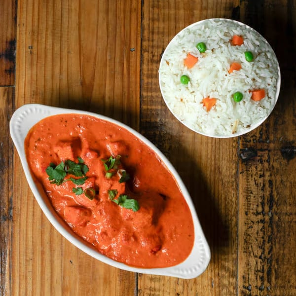 Indian butter chicken in a white dish with a side of rice and vegetables on a wooden table.